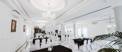 A spacious banquet hall with white walls, chandeliers, and a marble floor. Tables are set with white chairs and black tablecloths.
