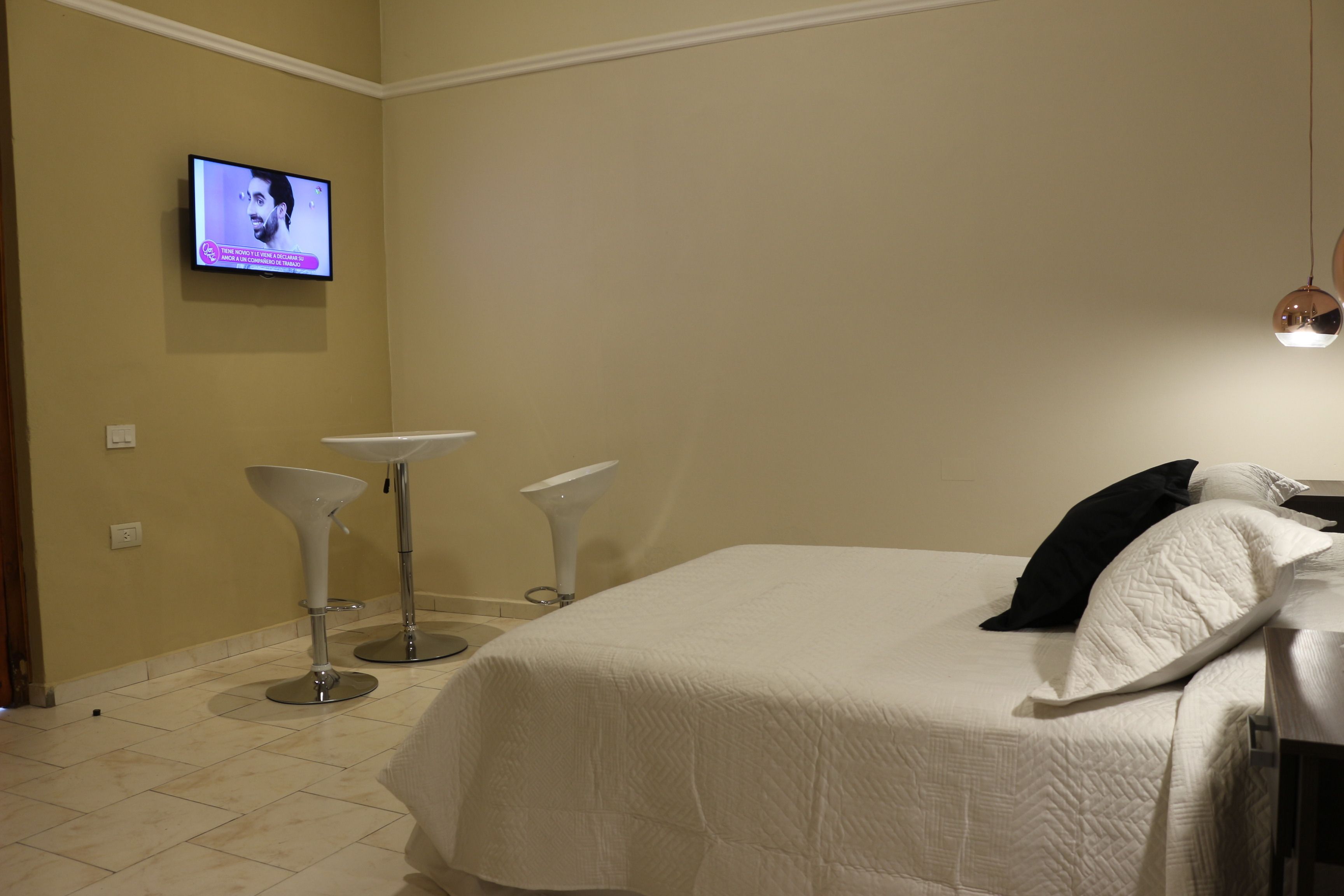 Une chambre d’hôtel avec un lit, un téléviseur fixé au mur et deux chaises blanches sur pied.
