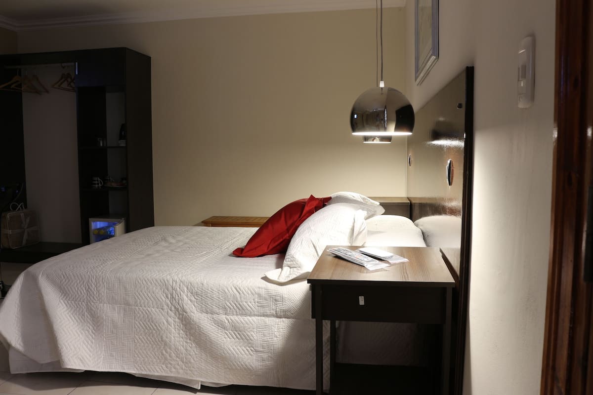 Una cama bien hecha con colcha blanca y almohada roja, una mesita de noche con un libro, y una lámpara colgante.