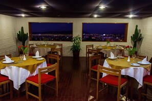 Un restaurante con mesas dispuestas para comer, amplias ventanas con vista a la ciudad y cactus como decoración.