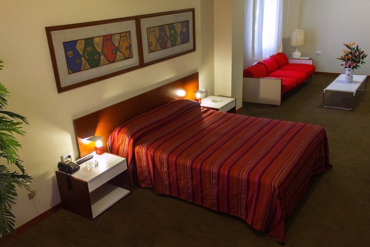 Ein Hotelzimmer mit Bett, Nachttisch, einer roten Couch, einem kleinen Tisch mit Lampe und zwei gerahmten Kunstwerken an der Wand.