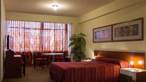 A hotel room with a bed, a desk, a chair, a small table, a plant, and two framed artworks on the wall. - Camino Real Hotel (Tacna)