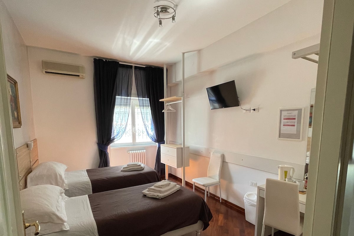A hotel room with two beds, a desk, a chair, a TV, and an air conditioning unit.