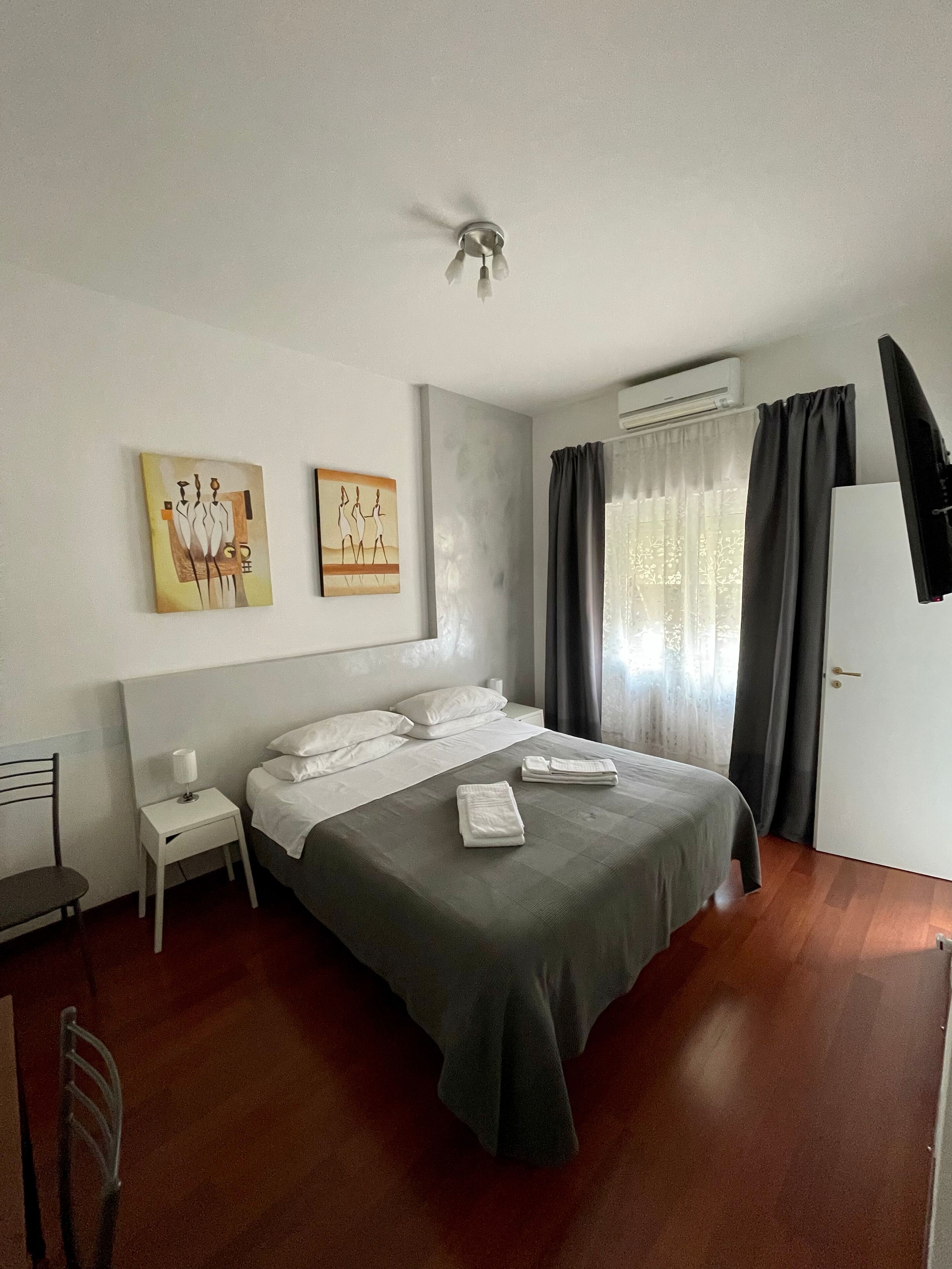 A hotel room with a bed, bedside tables, a chair, artwork on the wall, an air conditioning unit, and a window with curtains.
