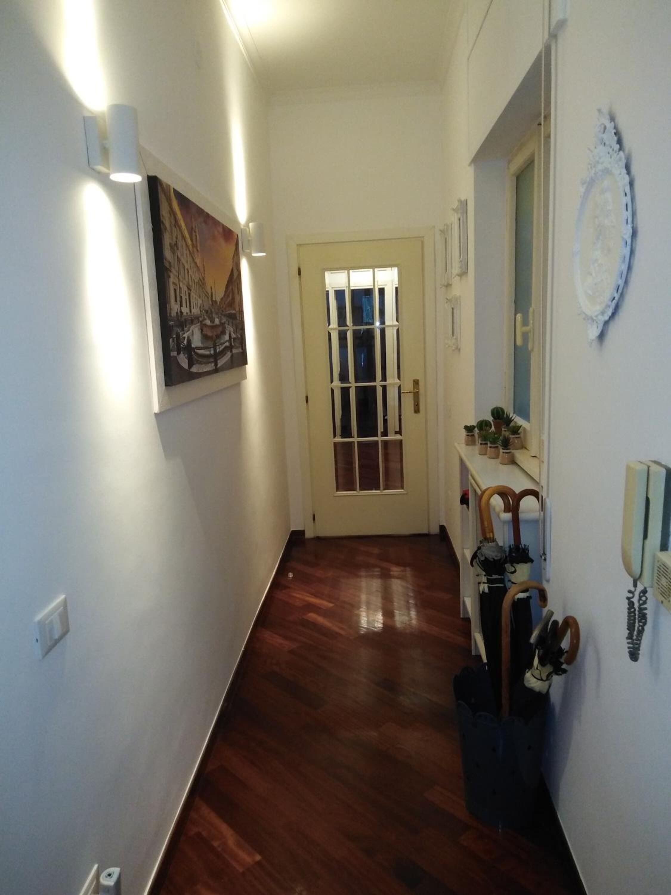 A hallway with a wooden floor, a door with a window, a wall-mounted telephone, and a wall-mounted light fixture.