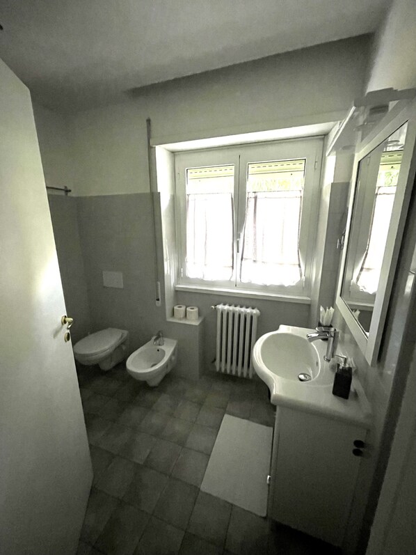 A bathroom with a toilet, bidet, sink, and a window with curtains. - Garden of Rome Guesthouse (Rome)