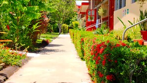A pathway flanked by lush greenery and red flowers leading to a building with red and beige exterior walls. - Villas Allen (Cahuita)