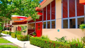 A building with a red and beige facade, multiple windows, and a well-maintained garden with red flowers and green hedges. - Villas Allen (Cahuita)
