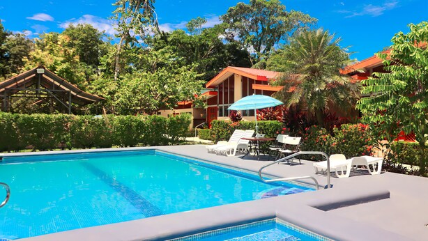 A swimming pool with lounge chairs and a covered seating area, surrounded by lush greenery and a building with a red roof. - Villas Allen (Cahuita)