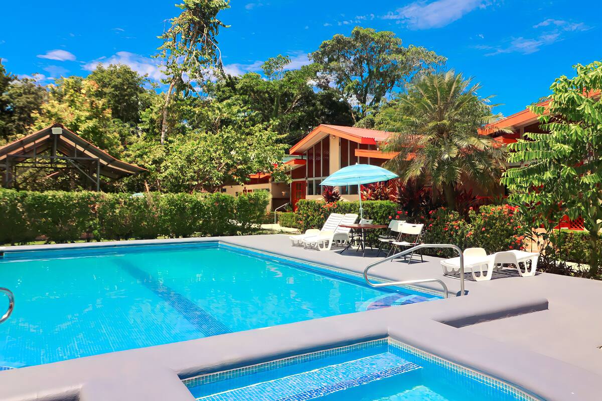 Ein Pool mit Liegestühlen und einer überdachten Sitzgruppe, umgeben von üppiger Vegetation und einem Gebäude mit rotem Dach.