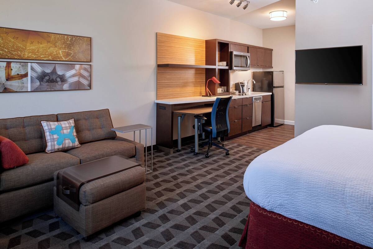 A compact hotel room with a kitchenette, a sofa with a pillow, a desk, and a bed.