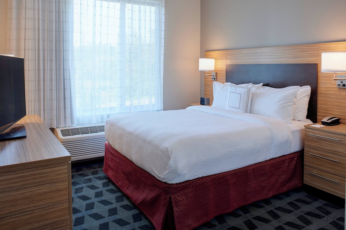 A hotel room with a large bed, a desk with a flat-screen TV, a bedside table with a lamp, and a window with blinds.