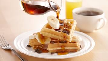 A plate of waffles with bananas and syrup, a glass of orange juice, and a cup of coffee on a wooden table.
