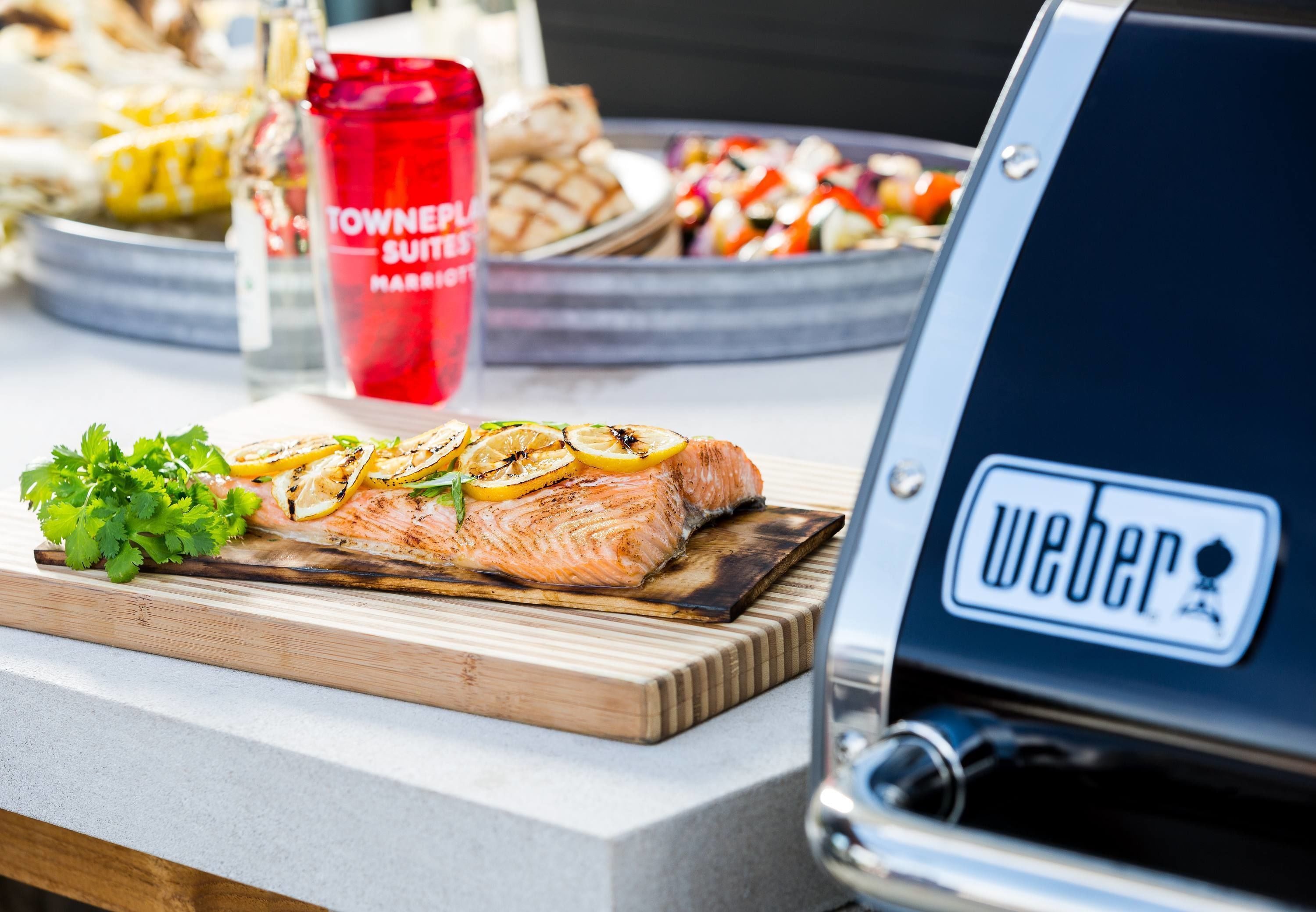 A grilled salmon on a wooden board with lemon slices and parsley, a red tumbler, and a Weber grill in the background.