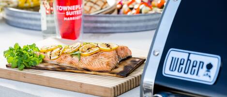 A grilled salmon on a wooden board with lemon slices and parsley, a red tumbler, and a Weber grill in the background.