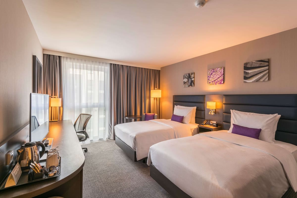 A hotel room with two beds, a desk, and a chair. There are lamps, a coffee maker, and a telephone on the desk.