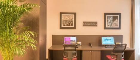 A modern office with two desks, each with a computer and a chair. There are framed pictures on the wall and a potted plant on the left.