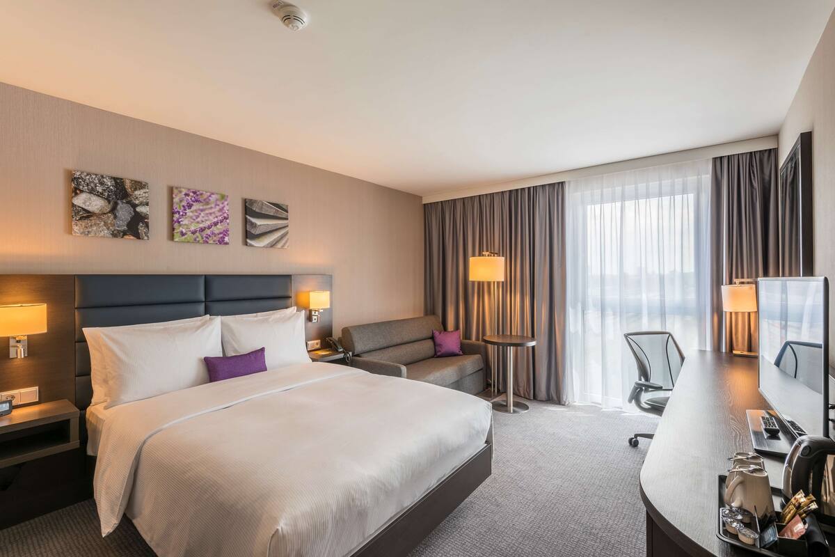 A hotel room with a large bed, a sofa, a desk with a computer, and a TV.
