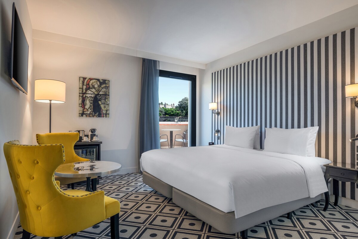 A hotel room with a bed, a yellow armchair, a small table, a painting, and a view of the outdoors.