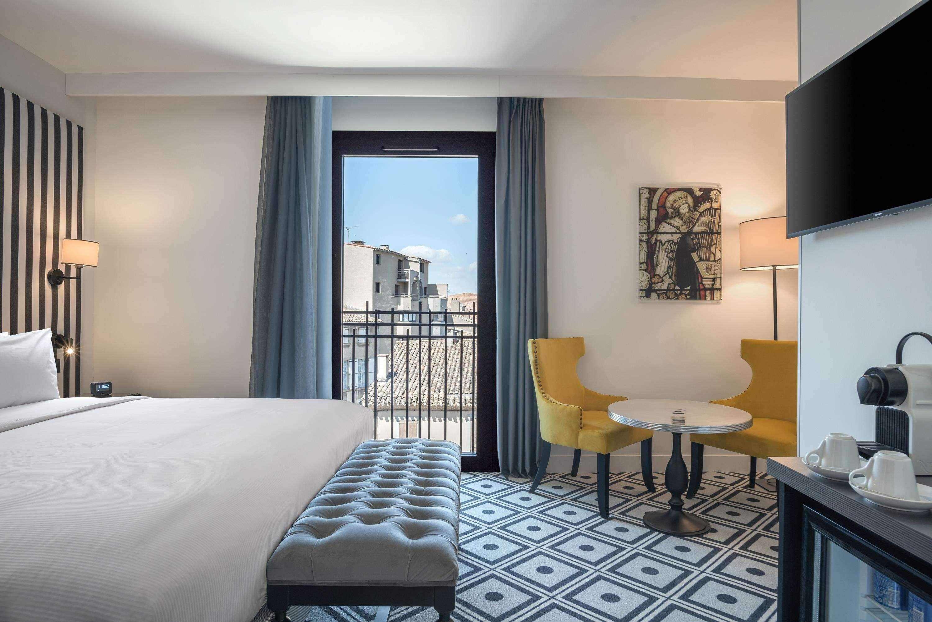 A hotel room with a bed, a bench, a small table with chairs, a TV, and a painting on the wall.