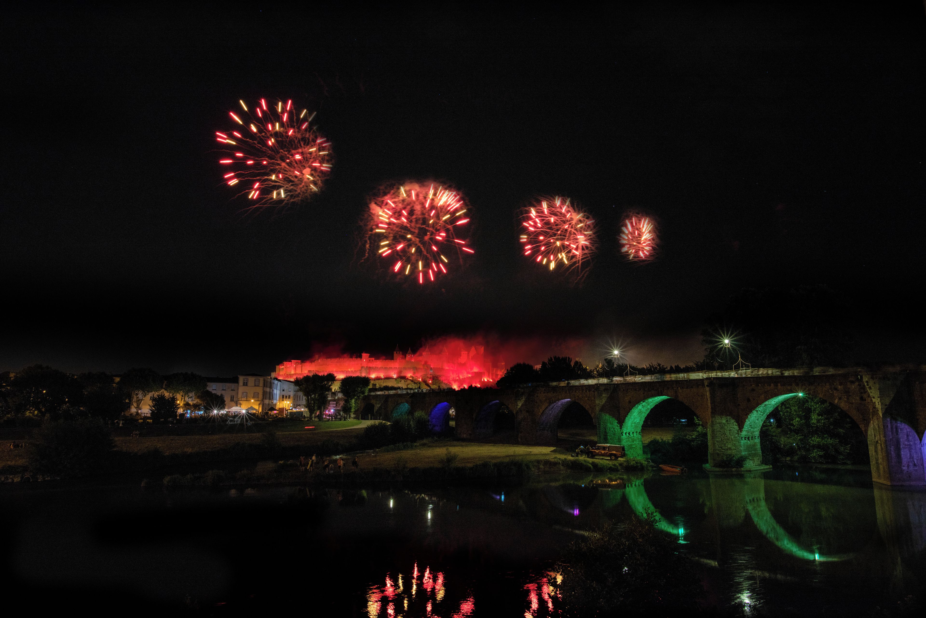 A nighttime view of a historic castle with fireworks in the sky, a bridge with green lighting, and a calm river reflecting the scene.