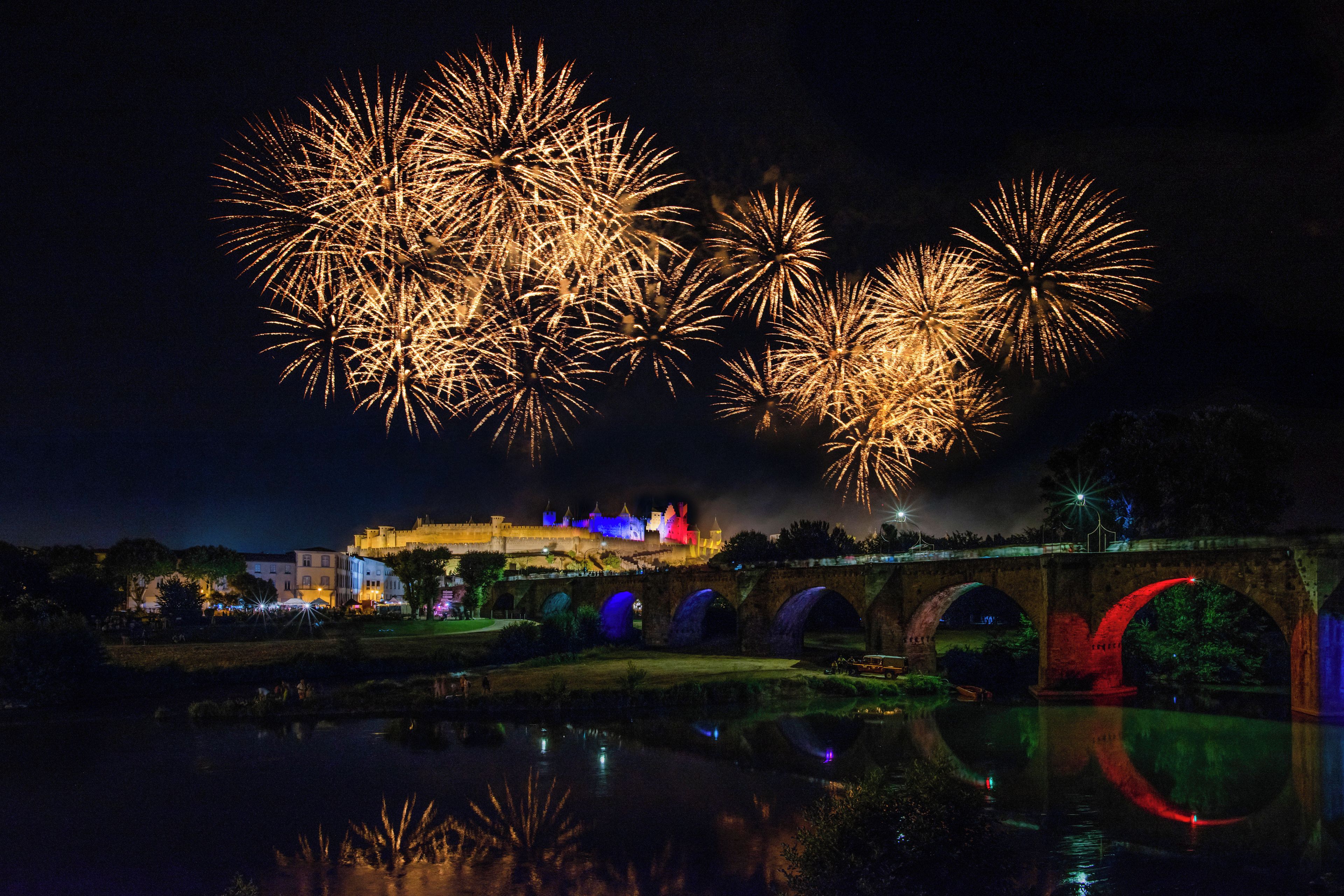 A nighttime view of a castle with fireworks in the sky, a bridge, and a body of water reflecting the scene.