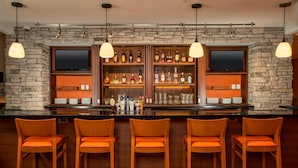 A bar area with a stone wall backdrop, wooden shelves with liquor bottles, and orange bar stools. - Fairfield Inn & Suites by Marriott Harrisburg International Airport (Middletown)
