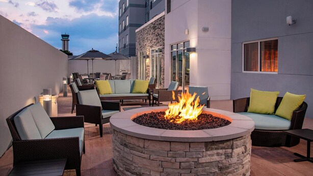 An outdoor seating area with a stone fire pit, cushioned chairs, and a view of a building with large windows and a tower. - Fairfield Inn & Suites by Marriott Harrisburg International Airport (Middletown)