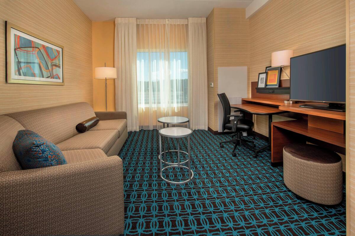 Une chambre d’hôtel avec un canapé, une petite table ronde, un bureau avec une chaise, une télévision et une moquette à motifs.