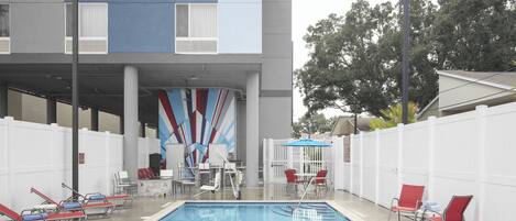 Una piscina con sillones de descanso y una zona de estar cubierta, rodeada por una valla blanca y un edificio de diseño moderno.