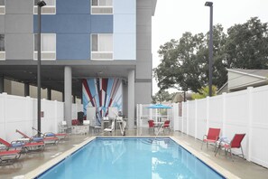 Una piscina con sillones de descanso y una zona de estar cubierta, rodeada por una valla blanca y un edificio de diseño moderno.