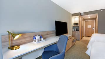 A hotel room with a desk, a blue chair, a television, a bed, and a kitchenette.