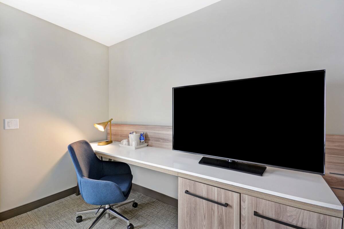 A hotel room with a large flat-screen TV on a white desk, a blue office chair, a desk lamp, and a wooden cabinet.