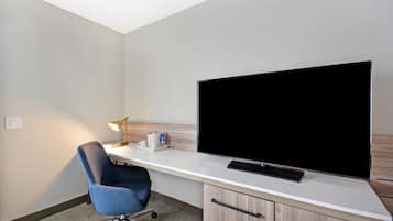 A hotel room with a large flat-screen TV on a white desk, a blue office chair, a desk lamp, and a wooden cabinet.