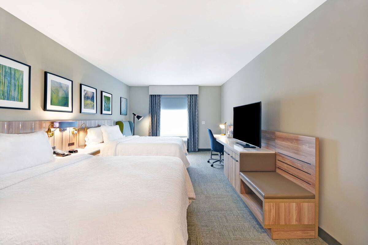 A hotel room with two beds, a desk, a TV, and framed artwork on the wall.