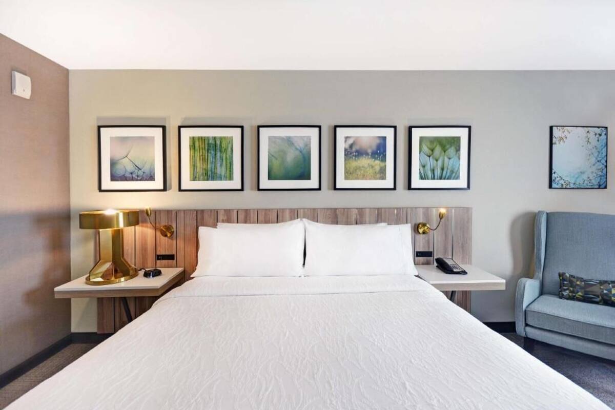 A hotel room with a large bed, a nightstand with a lamp, a chair, and five framed artworks on the wall.