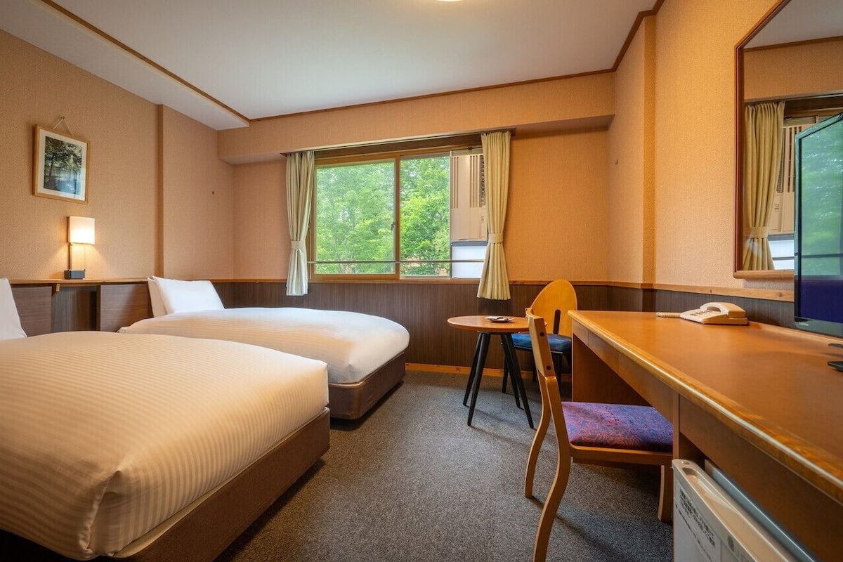 Hotellihuone, jossa on kaksi sänkyä, työpöytä, tuoli, televisio ja ikkuna, jossa on verhot.