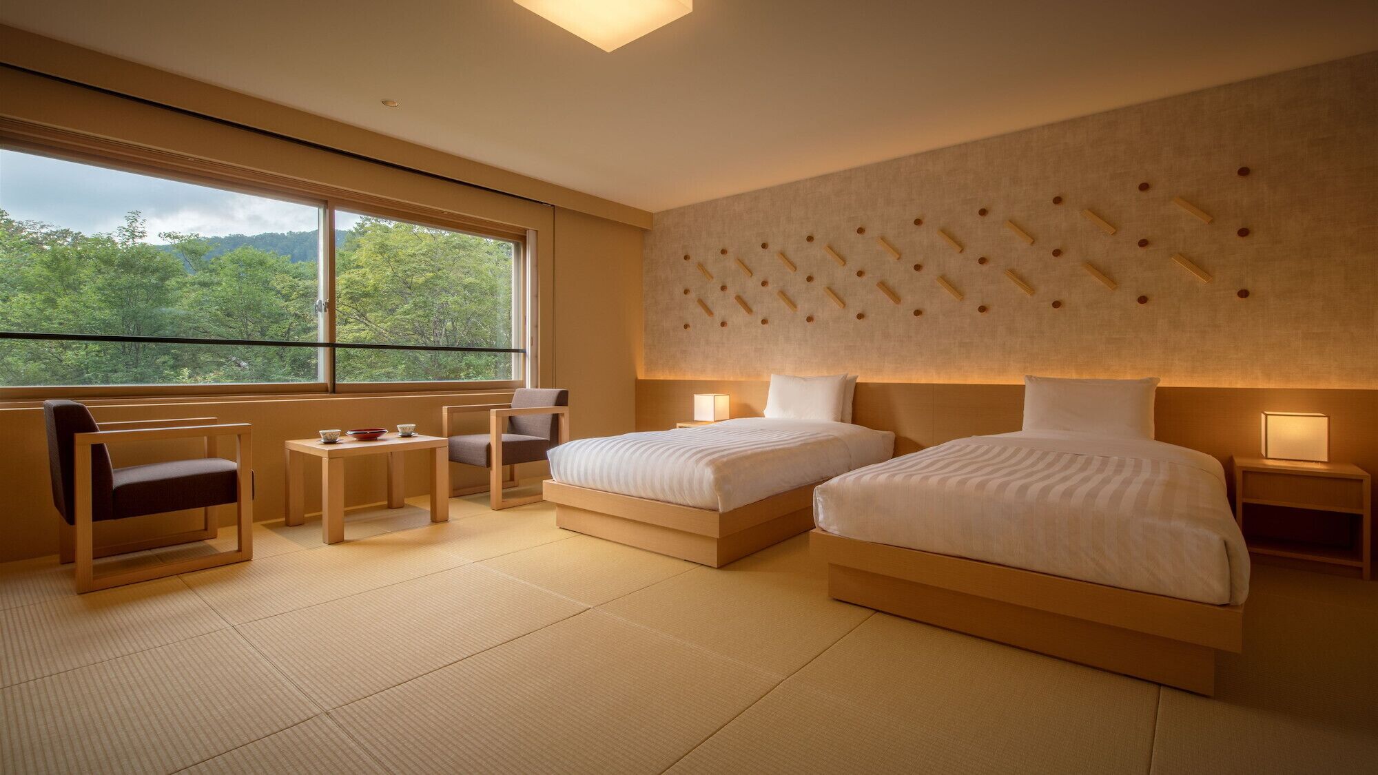 A hotel room with two beds, a sitting area with a table and chairs, a large window with a view of trees, and a textured wall design.