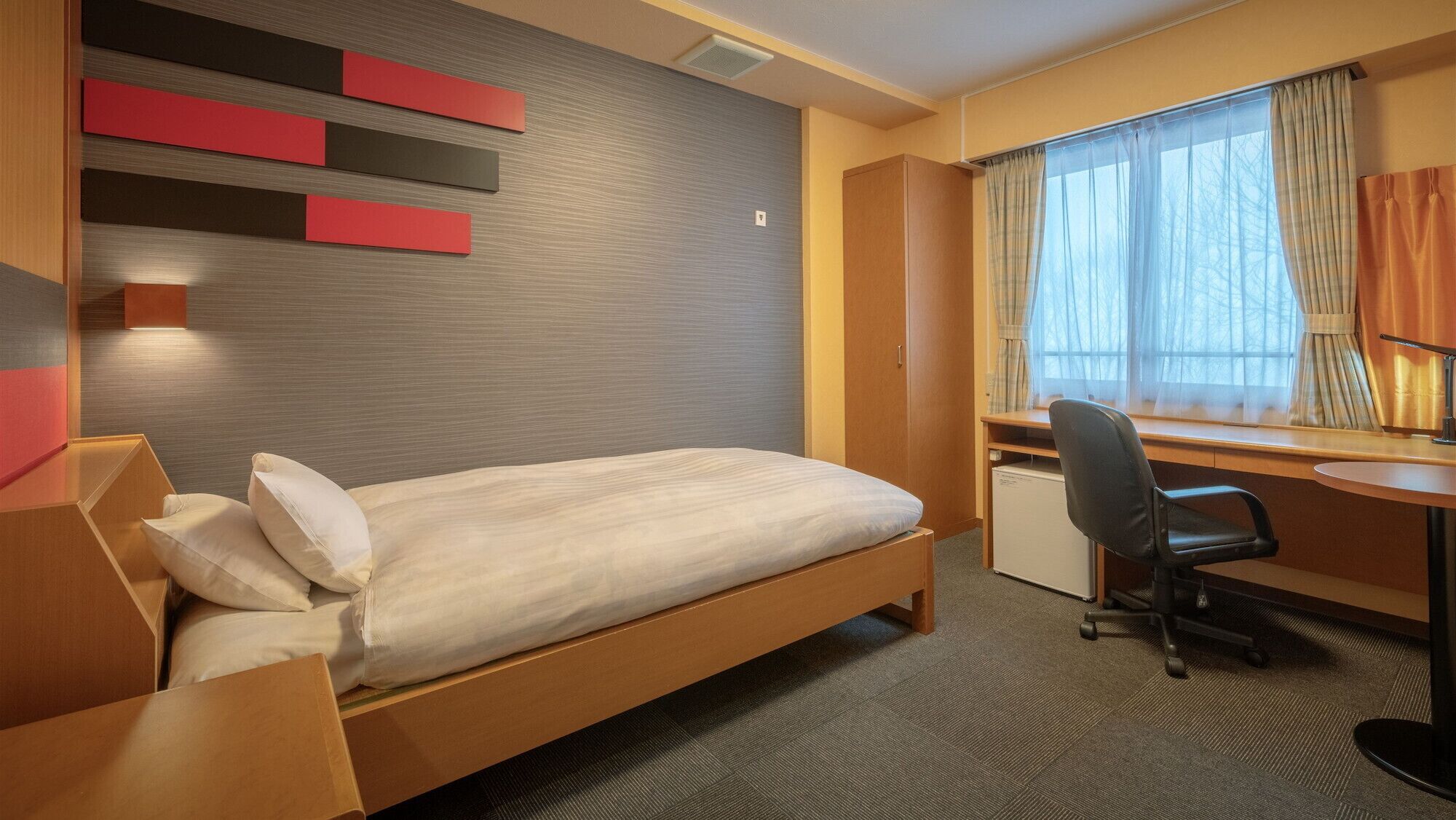 Habitació d'hotel amb llit, escriptori, cadira i finestra amb cortines.