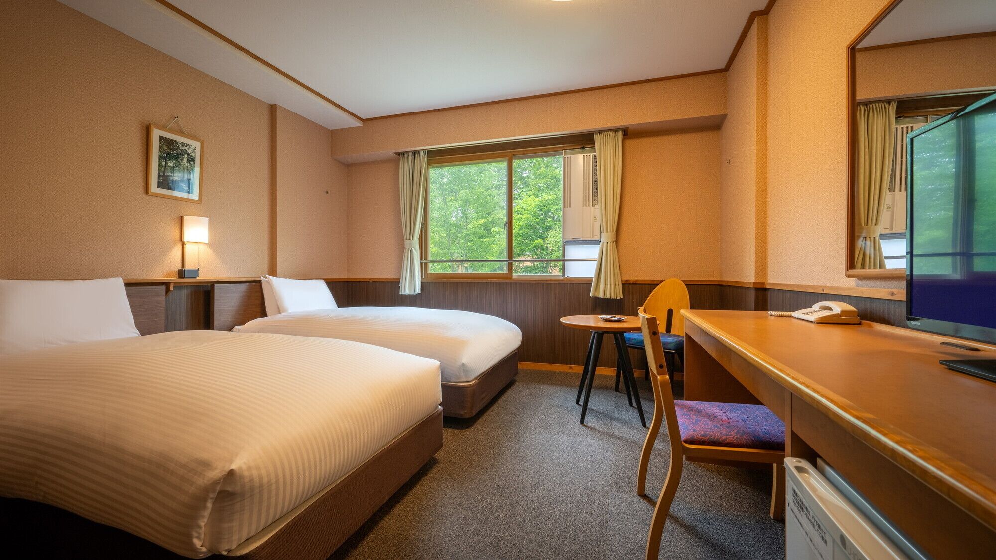 A hotel room with two beds, a desk with a chair, a window with curtains, a bedside lamp, and a framed picture on the wall.