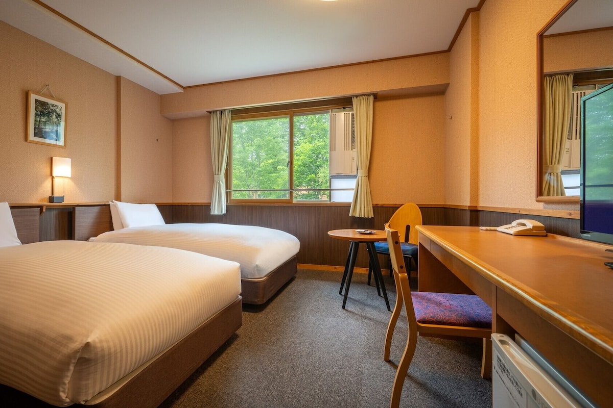 Hotellihuone, jossa on kaksi sänkyä, työpöytä tuolilla, ikkuna verhoilla, yöpöytälamppu ja seinällä oleva kehystetty kuva.