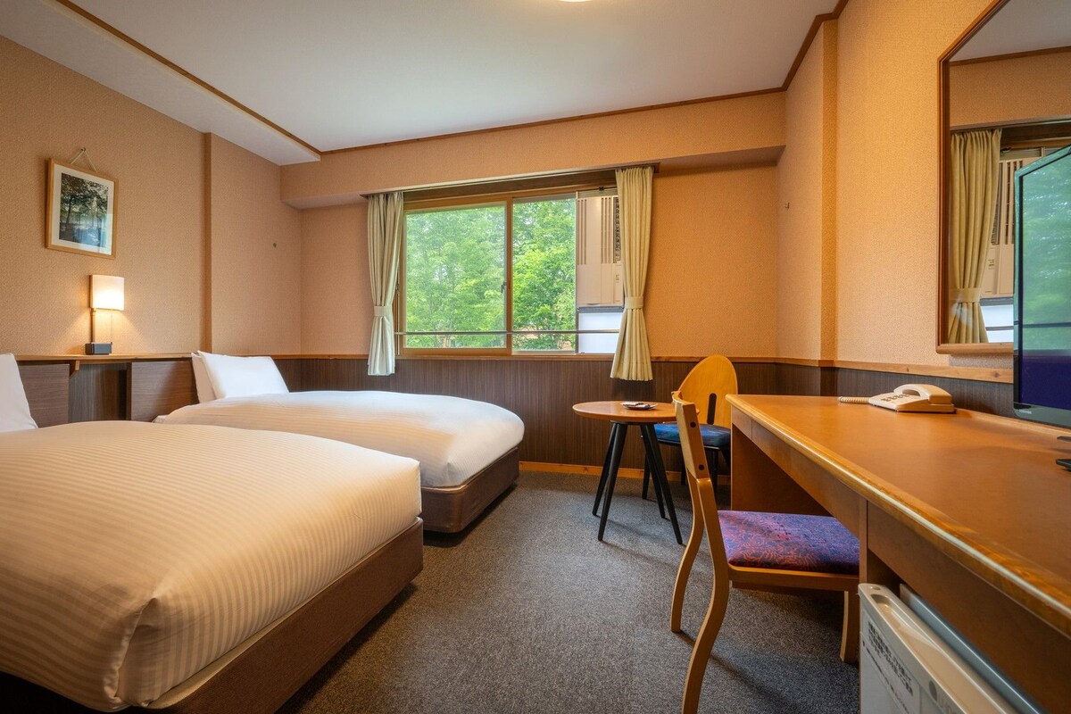Een hotelkamer met twee bedden, een bureau met stoel, een raam met gordijnen, een bedlampje en een lijst aan de muur.