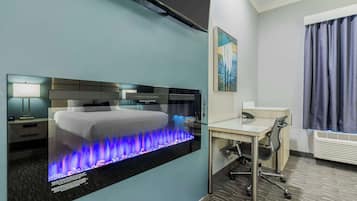 Habitación de hotel con cama, escritorio, silla, televisor montado en la pared y chimenea con llamas azules.