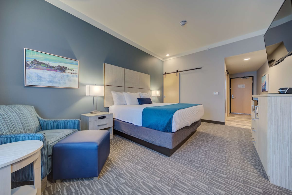 Une chambre d’hôtel moderne dotée d’un grand lit, d’un coin salon avec un canapé et un pouf, d’un bureau et d’un tableau au mur.