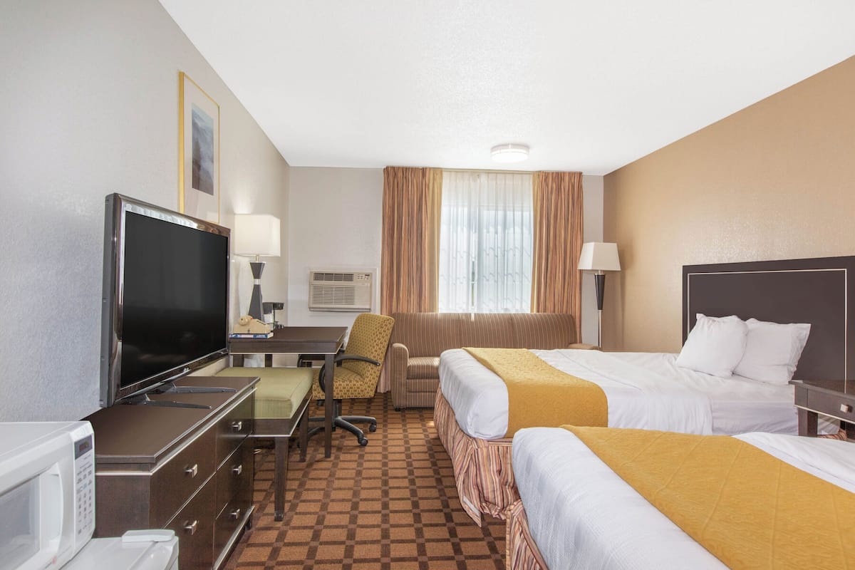 Ein Hotelzimmer mit zwei Betten, einem Schreibtisch mit Fernseher, einem Sessel, einer Mikrowelle und einem Fenster mit Vorhängen.