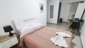En pænt redt seng med et rosa sengetæppe, en hvid pude og et hvidt håndklæde på sengen. Der er en sort lampe på et trænatbord ved siden af sengen.