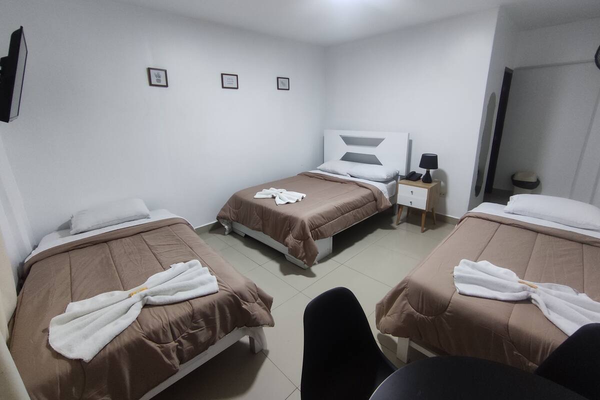 A hotel room with two single beds, each with a brown bedspread and a white towel. There is a small bedside table with a lamp and a phone. The room has white walls and a tiled floor.