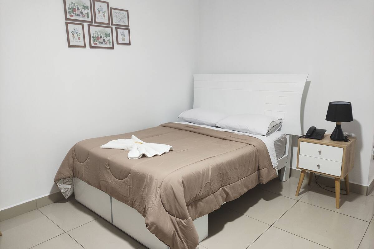 A neatly made bed with a brown bedspread and white pillows, a bedside table with a lamp and phone, and a wall with framed botanical illustrations.