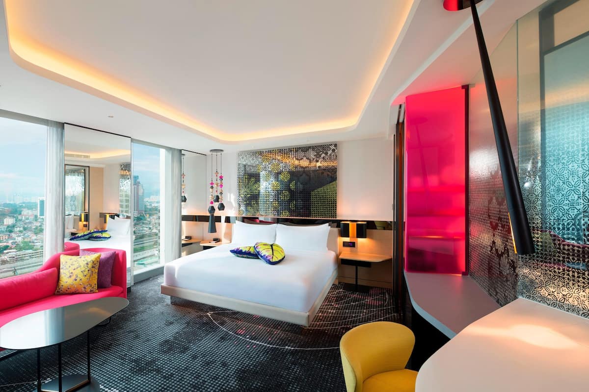 A modern hotel room with a large bed, a red sofa, a yellow armchair, and a city view.