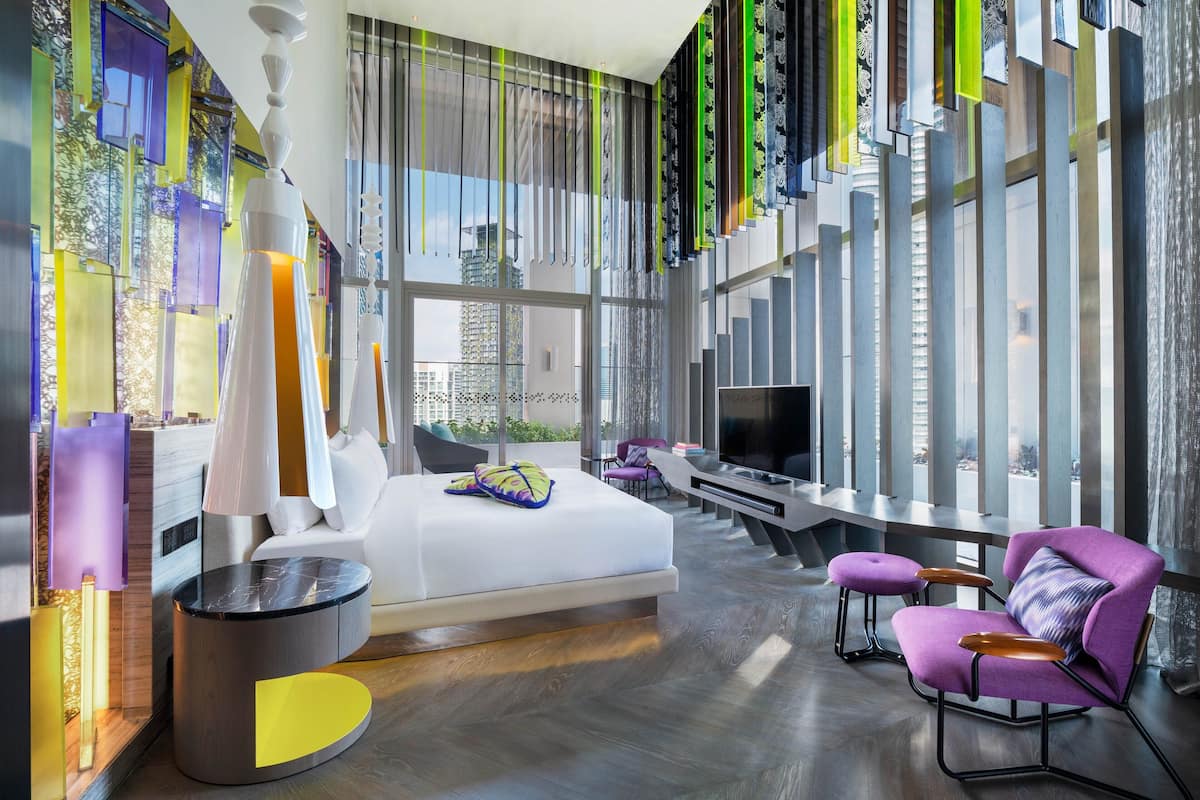 A modern hotel room with a large bed, a TV, and a seating area with a purple chair and ottoman.